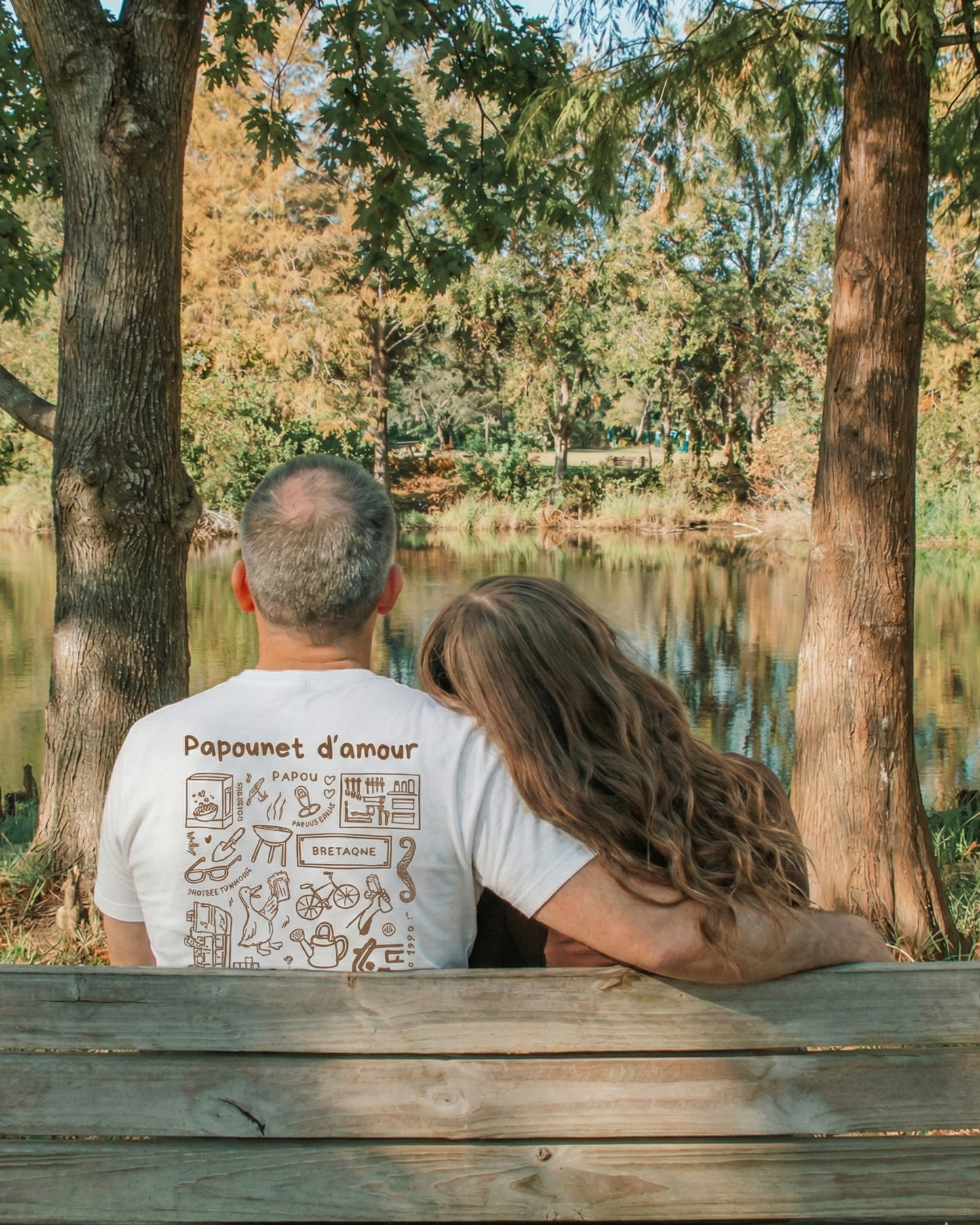 Photo d'un père et sa fille de dos sur un banc devant un lac. Le père porte le modèle de t shirt Archives de Koeur Indigo avec les souvenirs en commun avec sa fille et le texte personnalisé "papounet d'amour".