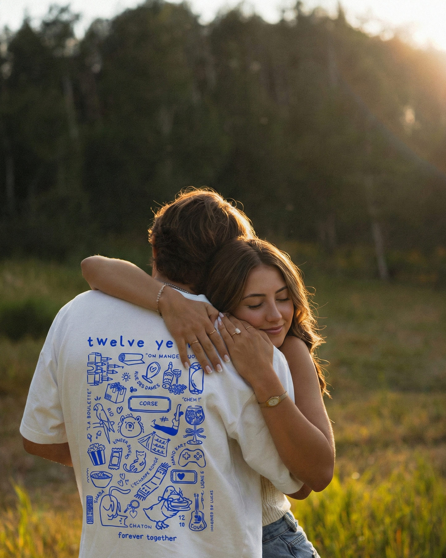 Photo d'un couple qui se prennent dans les bras dans un champ. L'homme porte le t shirt Archives de la marque Koeur Indigo avec les souvenirs en commun avec sa copine et les textes personnalisés "Twelve years" et "Forever together".